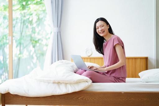 ベッドでパソコン中に笑うカメラ目線の女性 女性,ノートパソコン,ベッドの写真素材