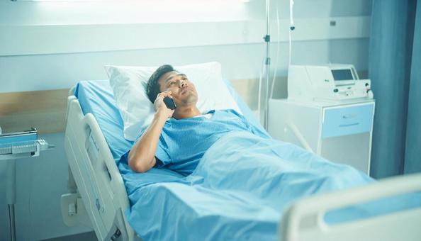 病室のベッドから携帯で話をする男性患者の写真