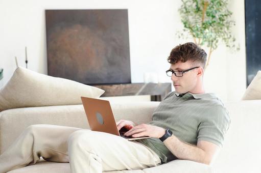 ソファに座って膝の上でノートパソコンを使っているメガネの男性 男性,外国人,欧米人の写真素材