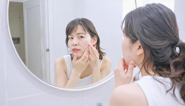 洗面所の鏡を見て頬の吹き出物に悩む女性 洗面所の鏡を見て頬の吹き出物に悩む女性の写真