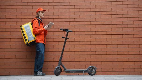 レンガ壁の前でタブレットを使うフードデリバリーの男性と電動キックボード 配達,フードデリバリー,デリバリーの写真素材