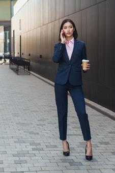 街なかでコーヒー片手に電話しているパンツスーツの女性 ビジネスウーマン,ビジネス,女性の写真素材