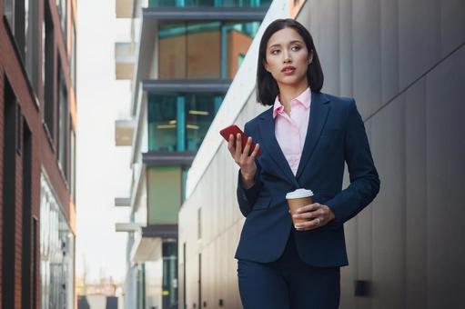街なかでコーヒーとスマートフォンを持って立つスーツの女性 ビジネスウーマン,ビジネス,女性の写真素材