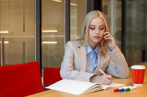 ノートにメモを取りながらスマートフォンで電話するビジネスウーマンの写真