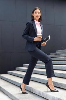 コーヒーとタブレットを持ってオフィスビルの外の階段を登っているパンツスーツのビジネスウーマン ビジネスウーマン,ビジネス,女性の写真素材
