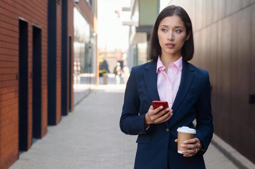 街なかでスマートフォンとコーヒーを持って立つビジネスウーマン ビジネスウーマン,ビジネス,女性の写真素材