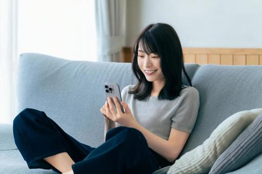 ソファに座ってスマホを使う笑顔の女性 ソファに座ってスマホを使う笑顔の女性の写真