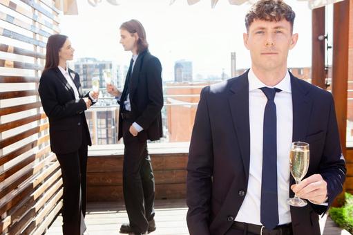 グラスを持ち微笑みカメラ目線の男性の後ろで会話中の男女の写真