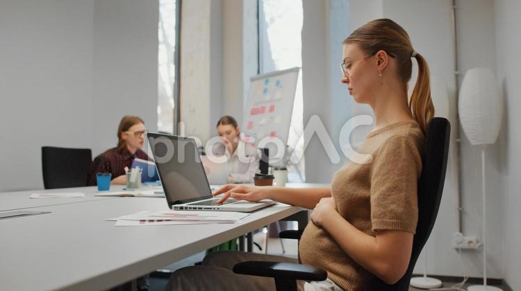 オフィスでお腹に手を置きながらノートパソコンを使っている妊娠中の女性 ビジネス,ビジネスマン,ビジネスウーマンの写真素材
