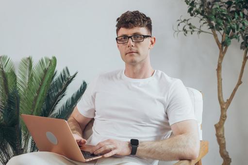 椅子に座って膝の上でノートパソコンを使うTシャツを着たカメラ目線の男性 男性,外国人,欧米人の写真素材