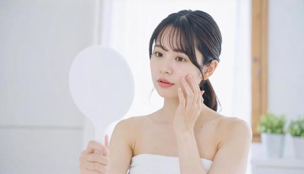手鏡で顔を見てお肌の悩みに悩む若い女性の写真