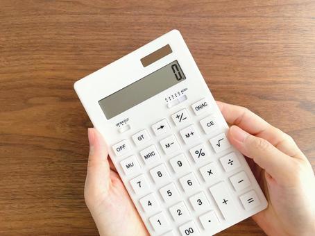 Woman's hand holding a white calculator on a wooden desk, JPG