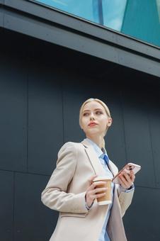 オフィスビルの外でコーヒーとスマートフォンを持って立っている女性 ビジネスウーマン,ビジネス,女性の写真素材