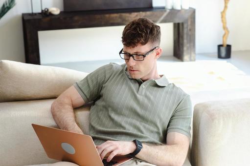 ソファに座って膝に乗せたノートパソコンを使う男性 男性,外国人,欧米人の写真素材
