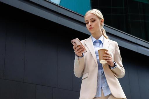 オフィスビルの外でコーヒー片手にスマートフォンを見ているビジネスウーマン ビジネスウーマン,ビジネス,女性の写真素材