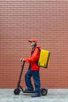 レンガ壁の前で電動キックボードに乗るフードデリバリーの男性 配達,フードデリバリー,デリバリーの写真素材