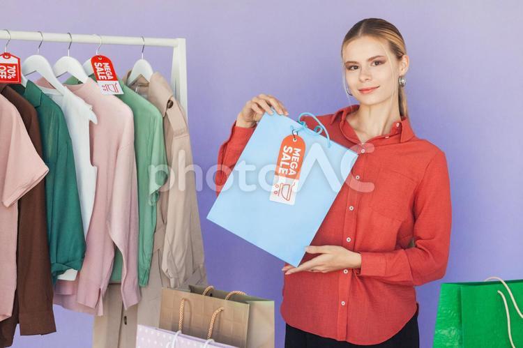 セールのタグの付いたハンガーに掛かった洋服と紙袋を持っているカメラ目線の白人女性 セール,ショップ,女性の写真素材