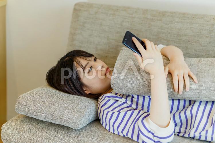 ソファーに寝ころんでスマホを見る女性 女性,ソファー,スマホの写真素材