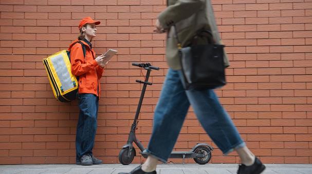 レンガ壁の前でタブレットを使うフードデリバリーの男性と通行人の女性 配達,フードデリバリー,デリバリーの写真素材