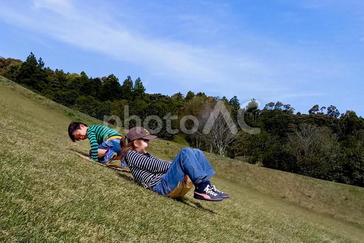 ＜素材＞公園の斜面で草スキーをする親子 子供,男性,女性の写真素材