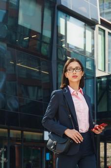 オフィスビルの外でスマートフォンを持って立つメガネを掛けたビジネスウーマン ビジネスウーマン,ビジネス,女性の写真素材
