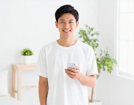 室内でスマホを持ち笑顔の白Tシャツの男性 室内でスマホを持ち笑顔の白Tシャツの男性の写真
