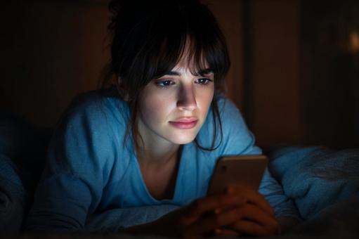 スマホを手にベッドに横たわる女性 スマホを手にベッドに横たわる女性の写真