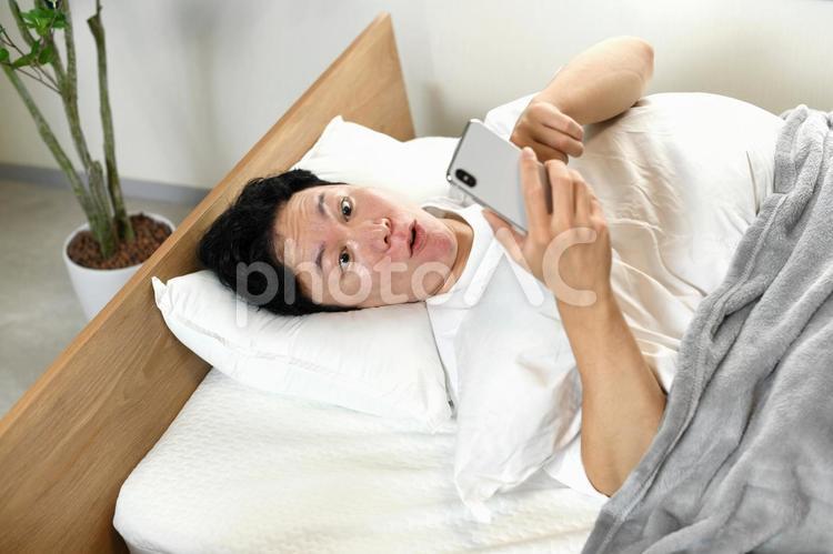 ベッドの上でスマホを持ち驚くアジア人男性 ベッド,男性,驚くの写真素材