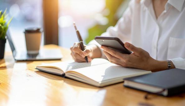 スマホ片手にノートに字を書く女性の手元 スマホ片手にノートに字を書く女性の手元の写真