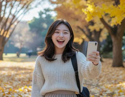 秋の公園でスマホを持ち笑顔を見せる女性 秋の公園でスマホを持ち笑顔を見せる女性の写真