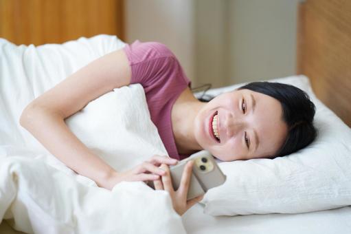 寝起きでスマホを操作する笑顔の女性 女性,スマートフォン,持つの写真素材