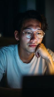 夜の部屋で手を頬に当てて思考する眼鏡男性の写真