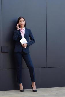 オフィスビルの黒い壁の前で電話するパンツスーツの女性 ビジネスウーマン,ビジネス,女性の写真素材