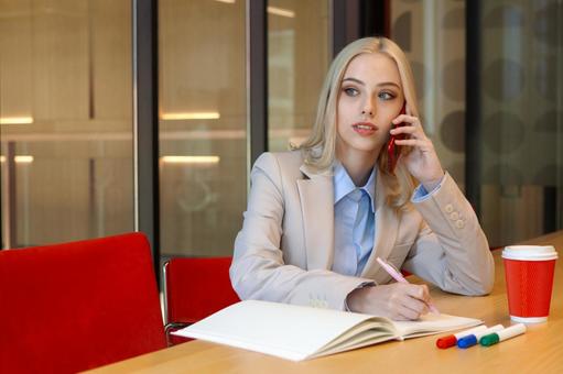 オフィスでノートにメモを取りながら電話している女性の写真