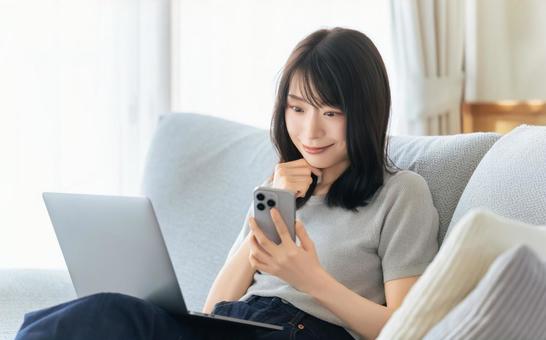 ソファに座ってスマホとパソコンを使う女性 ソファに座ってスマホとパソコンを使う女性の写真