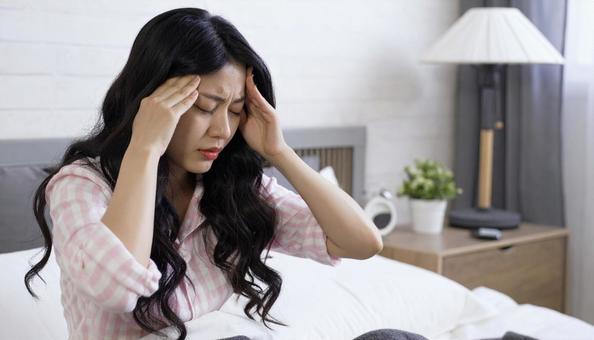 体調不良でベッドで頭を押さえる日本人女性の写真
