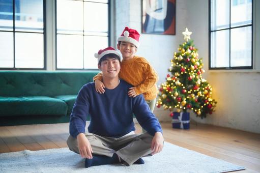 絨毯に座る男性の後ろで肩を持つ男の子とクリスマスツリー 絨毯に座る男性の後ろで肩を持つ男の子とクリスマスツリー 親子,男の子,小学生の写真素材