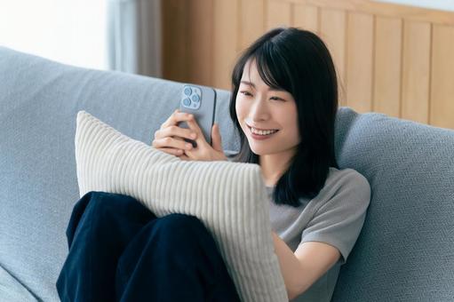 ソファに座ってスマホを使う笑顔の女性 ソファに座ってスマホを使う笑顔の女性 女性,スマホ,スマートフォンの写真素材