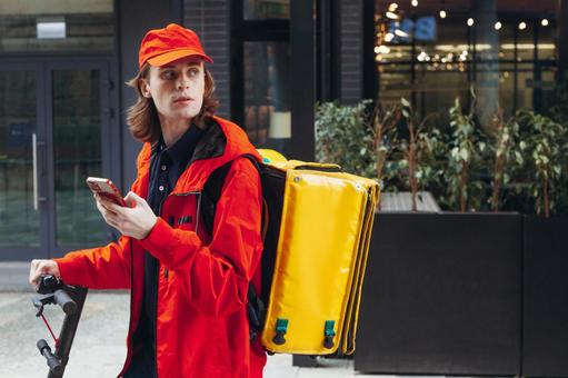 街なかでスマートフォンを見ている電動キックボードに乗る配達員の男性の写真