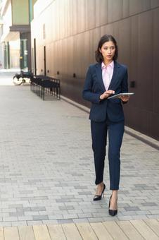 街なかでタブレットを持って歩くパンツスーツのアジア人女性 ビジネスウーマン,ビジネス,女性の写真素材