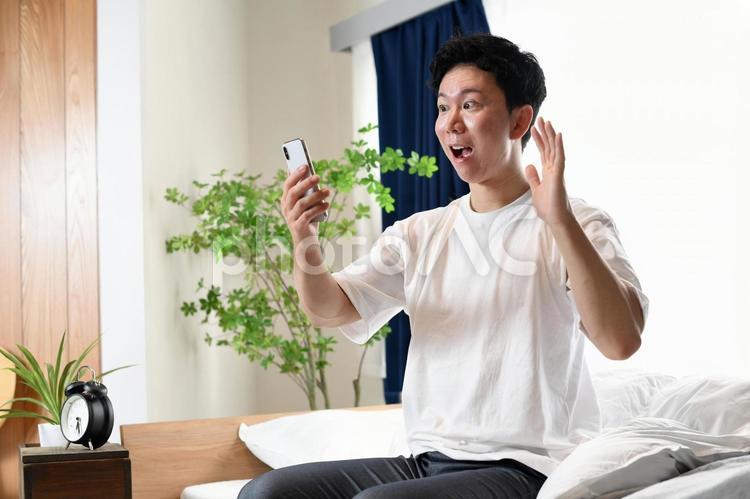 ベッドに座りスマホを持ち驚くアジア人男性 男性,驚く,スマートフォンの写真素材