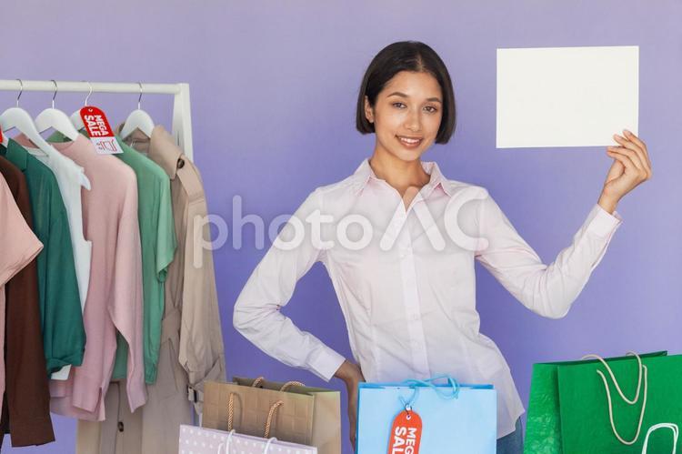 セールのタグの付いたハンガーに掛かった洋服の前で白い紙を見せるアジア人女性 セール,ショップ,女性の写真素材