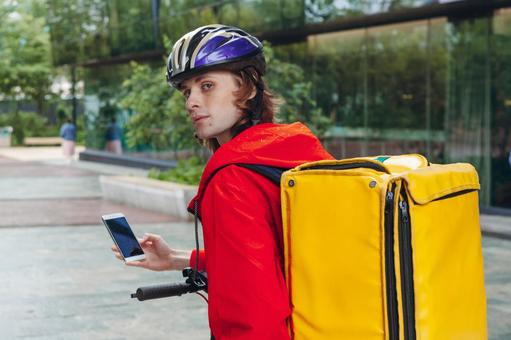 街なかでスマートフォンを見ているヘルメットを被ったフードデリバリーの男性 配達,フードデリバリー,デリバリーの写真素材