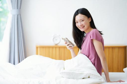 ベッドでスマホを持ち振り向く笑顔の女性 女性,スマートフォン,持つの写真素材