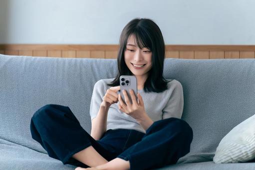 ソファに座ってスマホを使う笑顔の女性 ソファに座ってスマホを使う笑顔の女性の写真