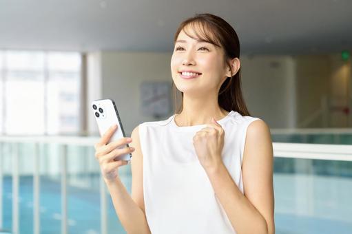 ジムでスマホを見てガッツポーズする女性 スポーツジム,女性,スマホの写真素材
