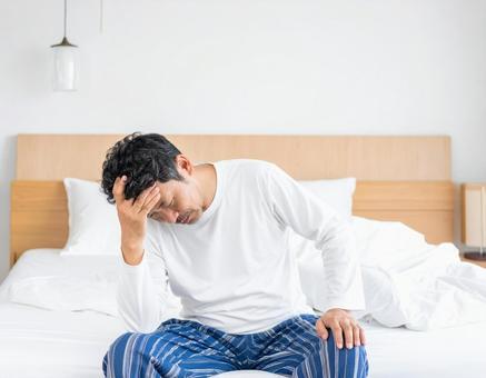 寝起きで座って頭を抱えるパジャマ姿の男性 寝起きで座って頭を抱えるパジャマ姿の男性の写真