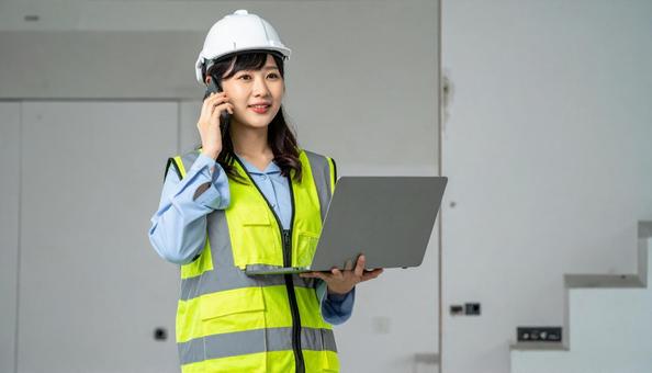 パソコンを抱えて電話する作業服の女性の写真