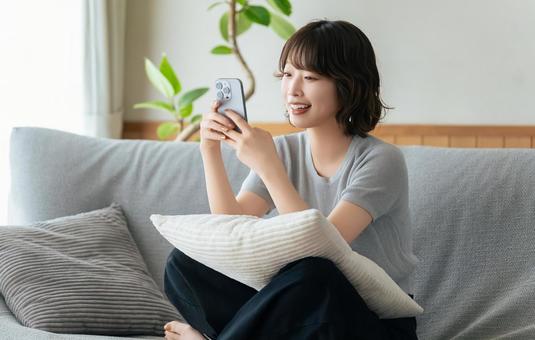 ソファに座ってスマホを使う笑顔の女性 ソファに座ってスマホを使う笑顔の女性 女性,スマホ,スマートフォンの写真素材