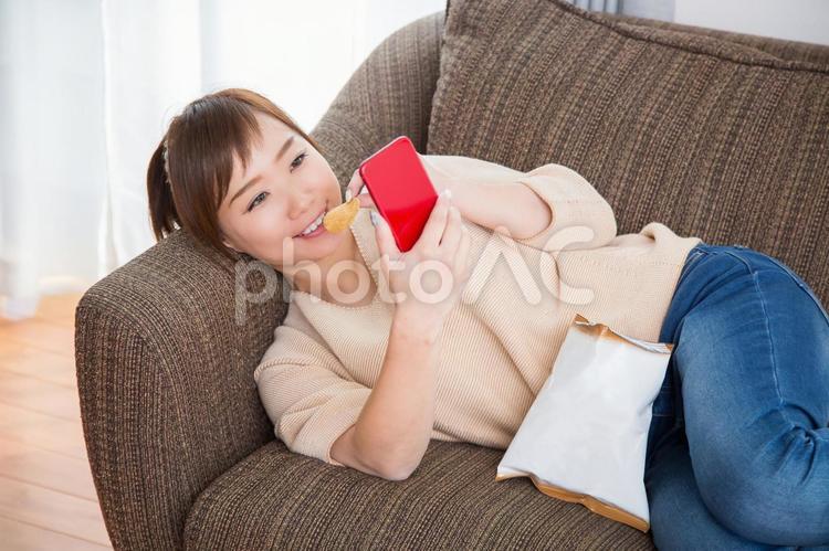 ソファでポテチを食べながらスマホを見る女性（笑顔） スマホ,女性,笑顔の写真素材